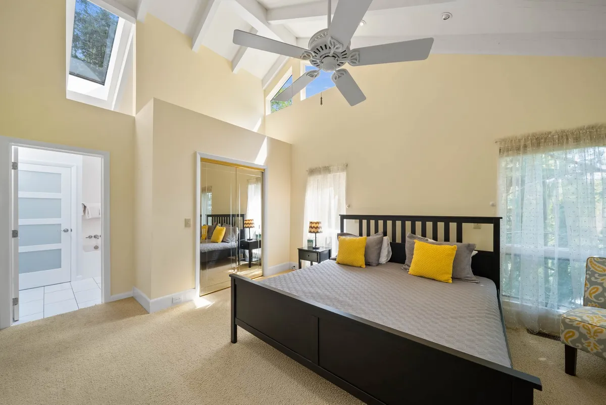 A calm bedroom featuring a cozy bed with yellow accents, large windows flooding the room with morning light, a ceiling fan, and a mirrored wardrobe. The setting is inviting and perfect for relaxing, reading, or enjoying a quiet moment.