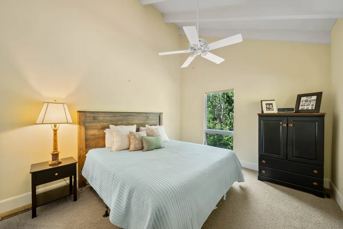 This image features a tranquil bedroom scene with a comfortable bed next to a large window, a warm lamp, and a dresser. The ambiance feels peaceful, inviting guests to relax and soak in the quietude of the mountain setting. The window suggests a connection to the outdoors, hinting at the natural surroundings just outside.