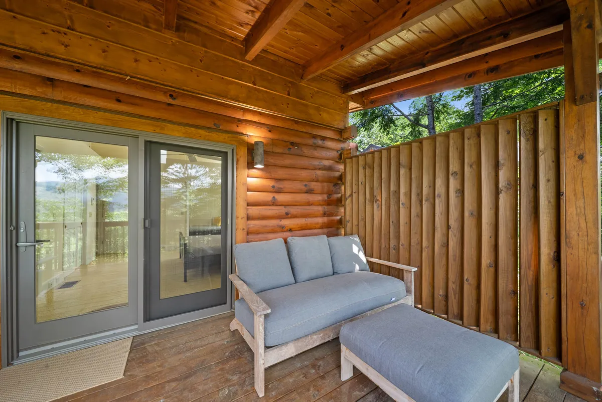 This image captures an inviting outdoor seating area, composed of comfortable chairs positioned on a wooden porch. Glass doors in the background provide a seamless transition between the cozy indoor space and the natural world outside. The tranquil scene is surrounded by lush greenery, evoking the serenity of the mountain environment.