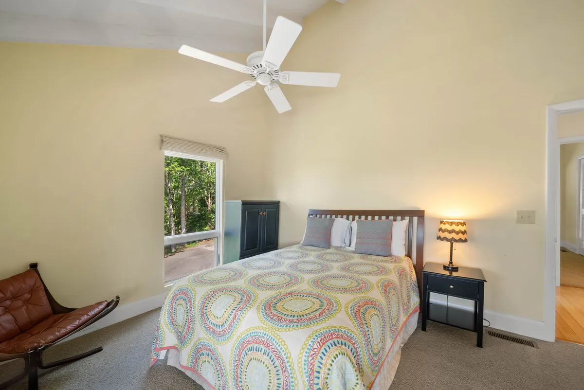A cozy bedroom with a patterned quilt on the bed, ceiling fan, window letting in soft daylight, and an armchair perfect for reading or morning coffee. The atmosphere is peaceful and welcoming, ideal for both quiet retreats and friendly gatherings.