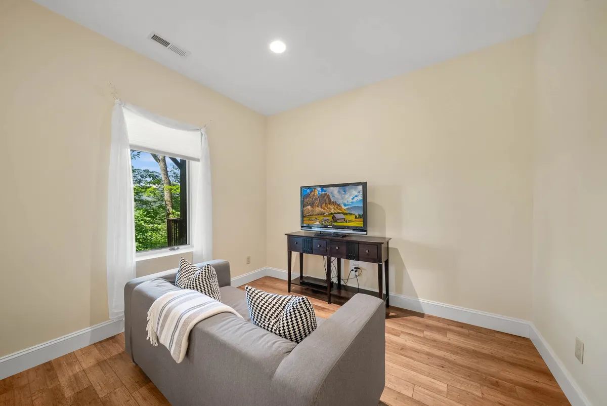 A cozy living room with a plush couch, a window framing the outdoors, and a side table. Though a TV is present, the focus of this image is the inviting setting, ideal for quiet reading, chatting, or simply gazing outside.