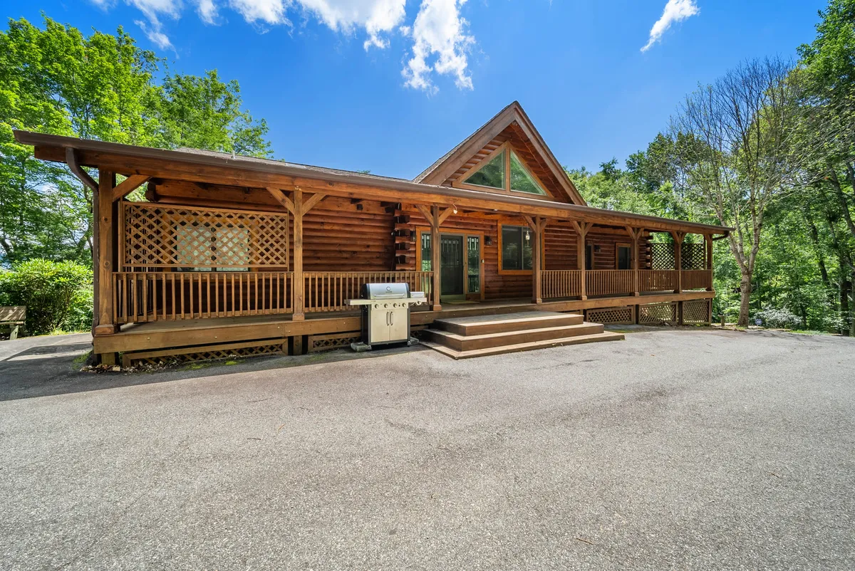 The image showcases a classic log cabin nestled in the mountains, featuring a spacious front porch equipped with comfortable seating and a gas grill. The porch overlooks a beautiful valley, bathed in the warm glow of sunset, creating an inviting atmosphere for outdoor dining, barbecuing, and relaxing while taking in the mountain views.