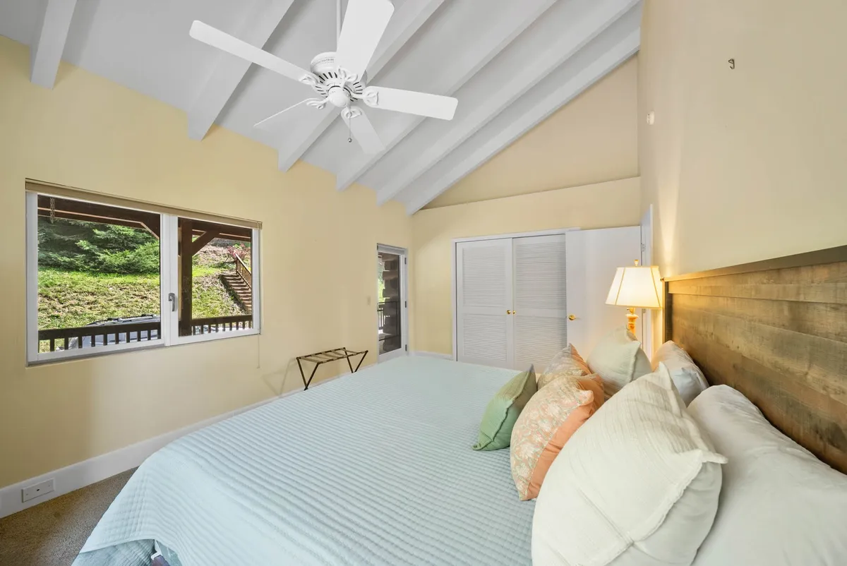 A calm, inviting bedroom featuring a large bed with decorative pillows, a wooden headboard, ceiling fan, and a generous window opening to mountain views. The gentle morning light suggests peaceful, extended stays and comfortable relaxation.