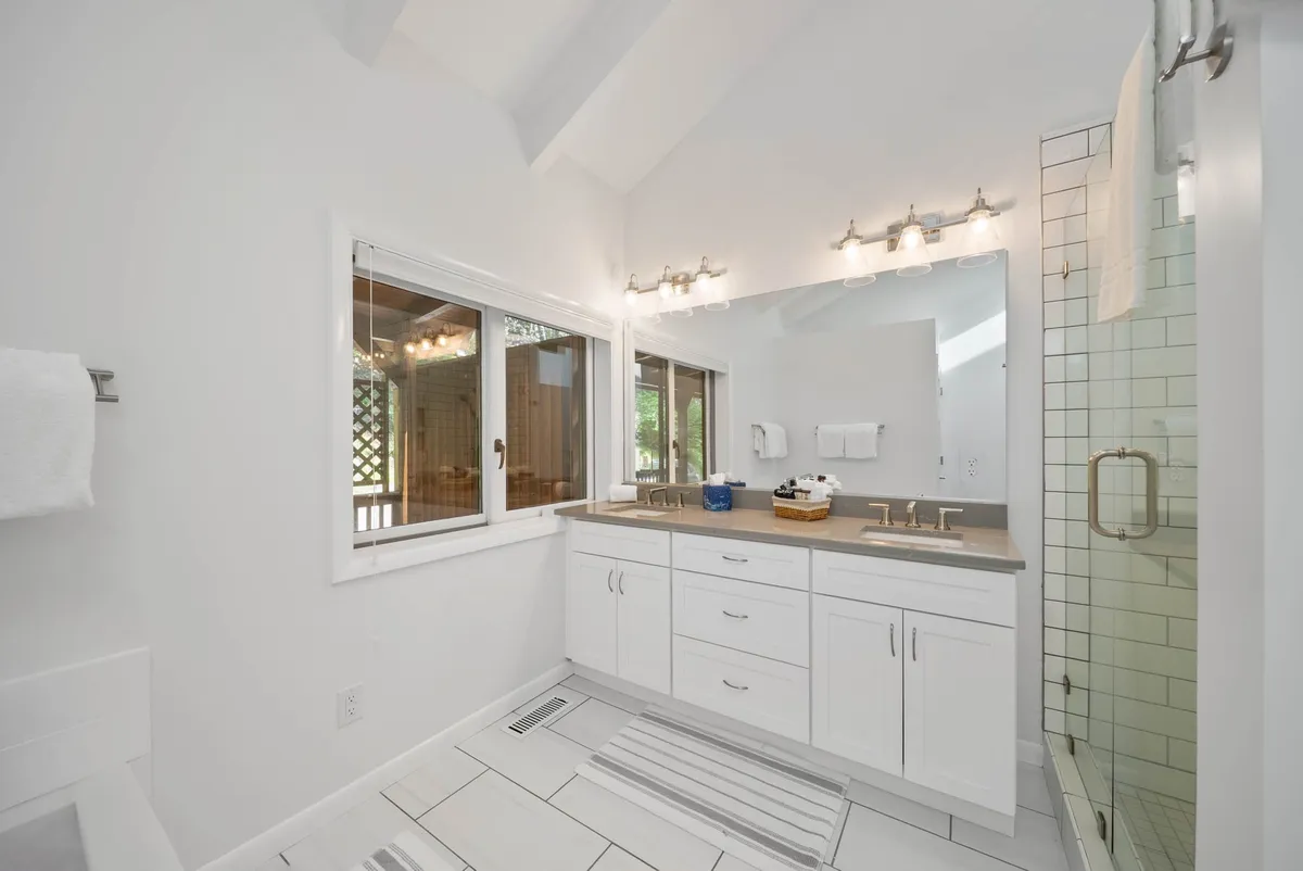 A modern bathroom with a double vanity, expansive mirror, glass shower enclosure, and a window with a tranquil view. The space is bright and serene, ideal for refreshing morning routines.