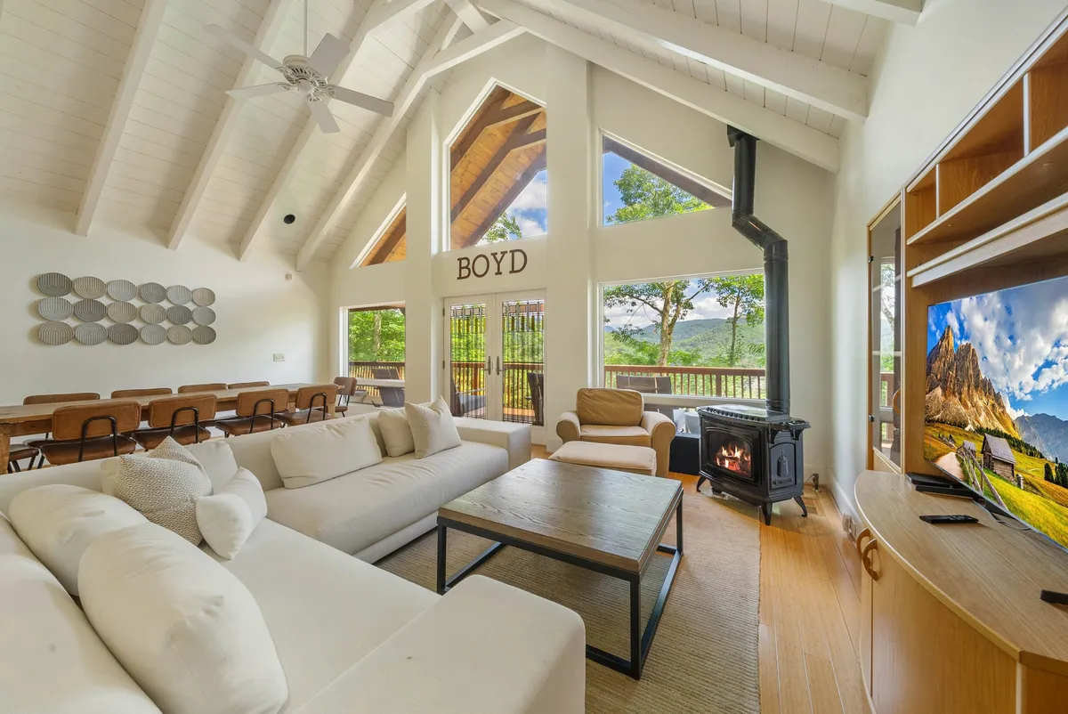 A spacious living room featuring a large sectional sofa, wooden coffee table, tall windows, and a prominent fireplace. The setup is ideal for relaxing with family, playing board games, or enjoying quiet moments by the fire, all while taking in natural views through the expansive windows.