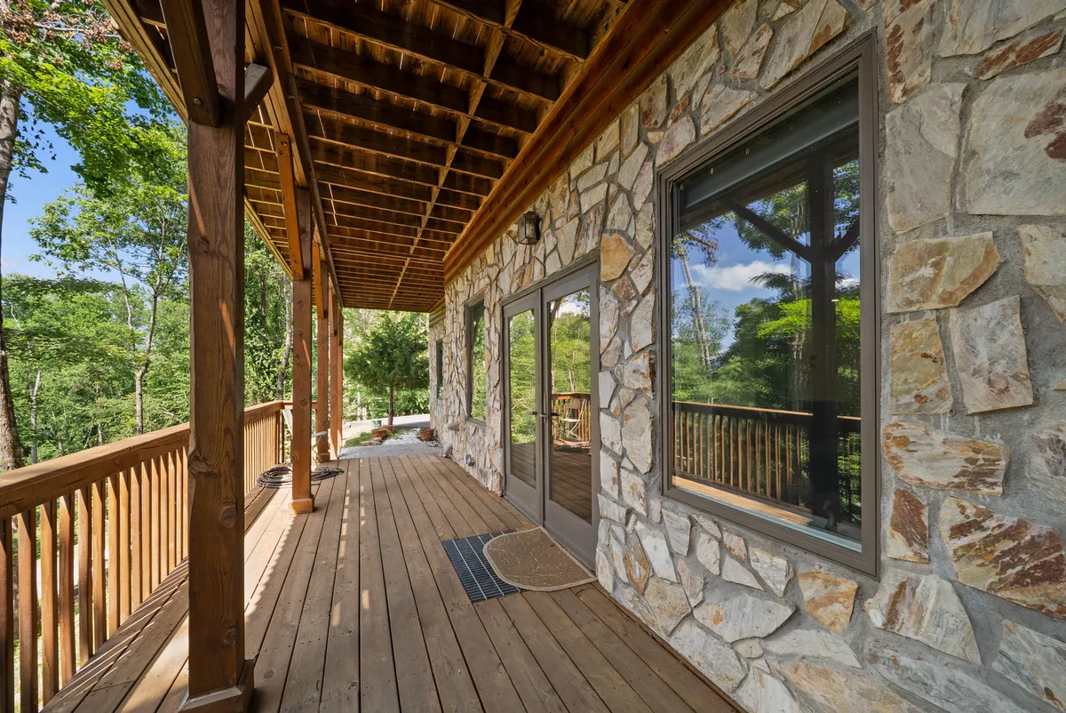 This image features the striking stone exterior of the home, a spacious wooden deck, large windows, and an overhanging roof. The setting radiates a welcoming ambiance and hints at a seamless connection between indoor comfort and outdoor exploration. The porch invites relaxation and social gatherings, with scenic views of the surrounding nature visible from the deck.