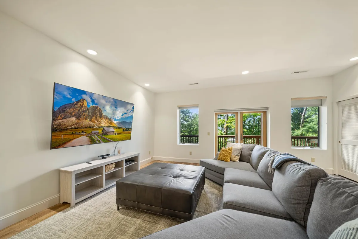 Another view of a cozy entertainment space, highlighting a large sectional sofa arranged for group seating, a television displaying a scenic landscape, and expansive windows that bring in natural light and connect the indoors to the outdoors. The setup encourages socializing and relaxation after a day of adventure.