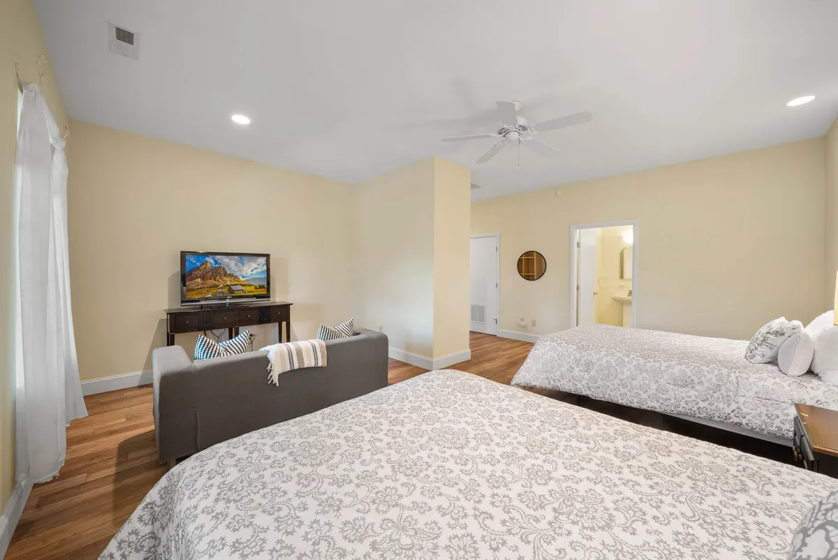 A spacious downstairs bedroom with two large beds adorned with decorative bedding, a modern TV on a wooden stand, a small sofa with accent cushions, and a round mirror. The setup provides ample space and privacy, ideal for families or groups.