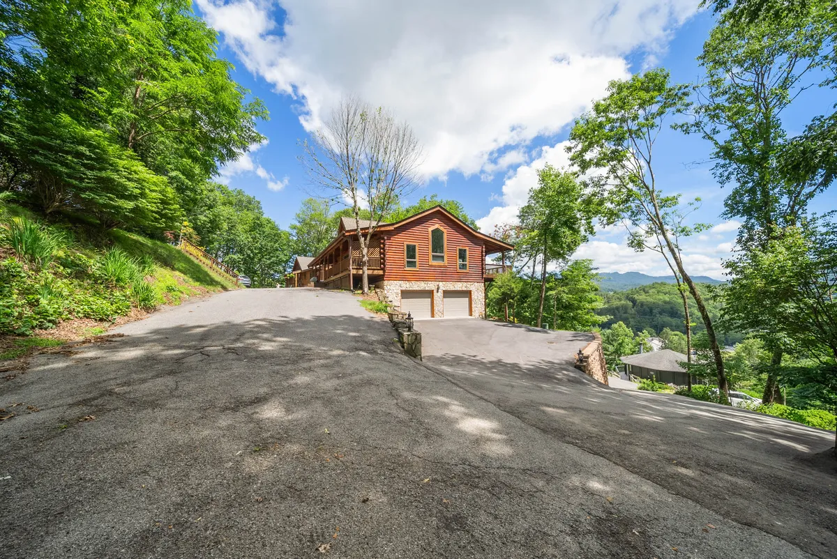 The image shows the home nestled among trees, with the driveway leading up to it and a sweeping mountain view in the background. The exterior feels serene and welcoming, framed by greenery that highlights the home's tranquil setting and accessibility to outdoor activities or quick trips into town.